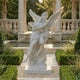 The Angelic Embrace Marble Outdoor Sculpture (260cm) by Giant Sculptures is displayed in a formal garden, surrounded by manicured bushes, stone columns, and a reflecting pool.