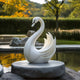 The Elegant Swan White Marble Outdoor Sculpture by Giant Sculptures is showcased elegantly on a round stone pedestal in a tranquil garden, with blurred greenery and distant yellow-leaved trees accentuating the serene outdoor scene.