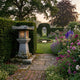 Large Japanese Temple Lantern 120cm by Giant Sculptures in carved grey granite, glowing softly on a brick path beside clipped hedges, lavender and foxgloves.