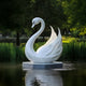 The Radiant Swan White Marble Outdoor Sculpture by Giant Sculptures stands gracefully in a serene pond surrounded by lush greenery, with water reflecting the sculpture and foliage, crafting a peaceful, nature-inspired art setting.