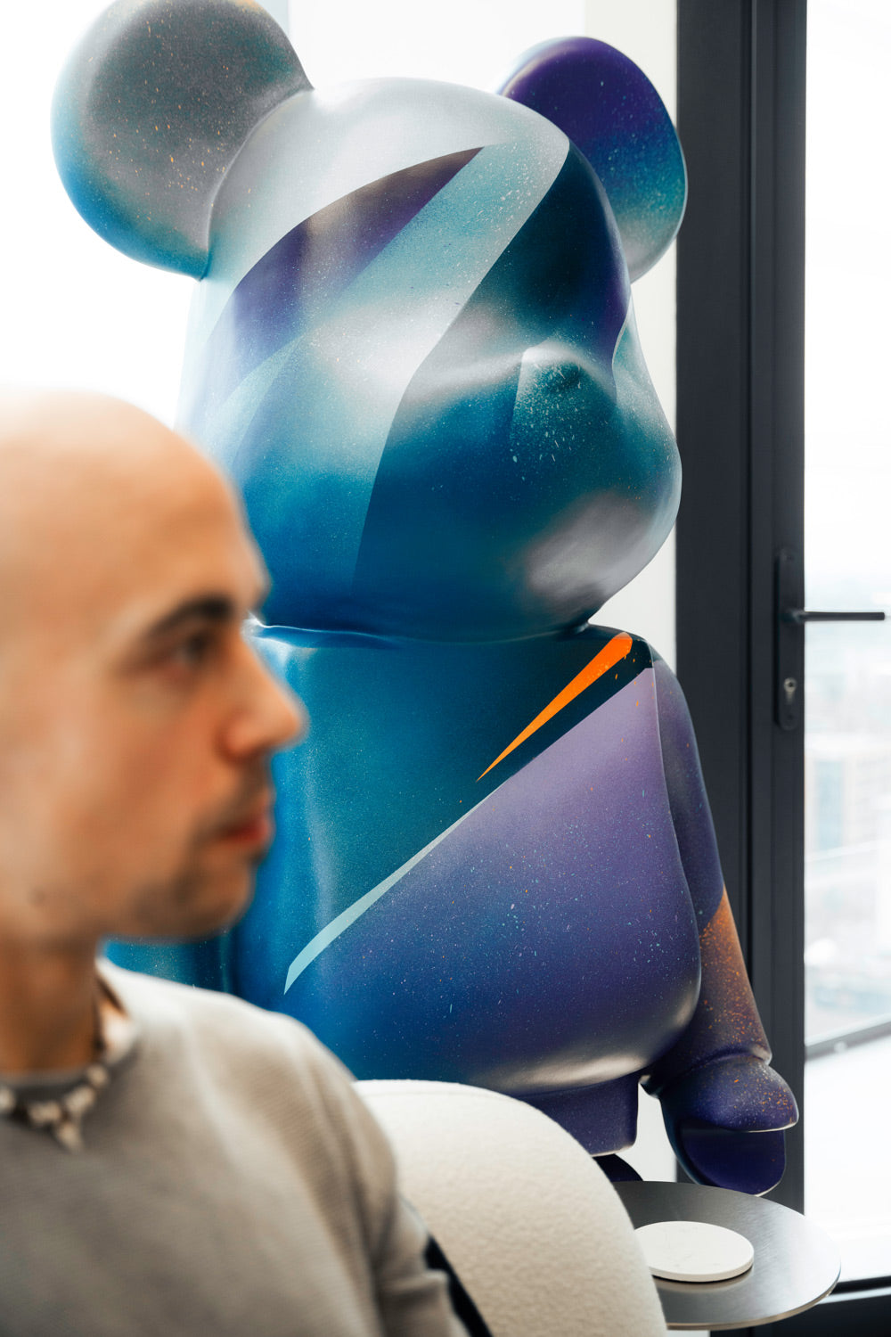 A bald person in focus sits indoors next to a large, colorful abstract teddy bear sculpture with a galaxy-like design. The sculpture has shades of blue, purple, and orange, and is positioned near a window.