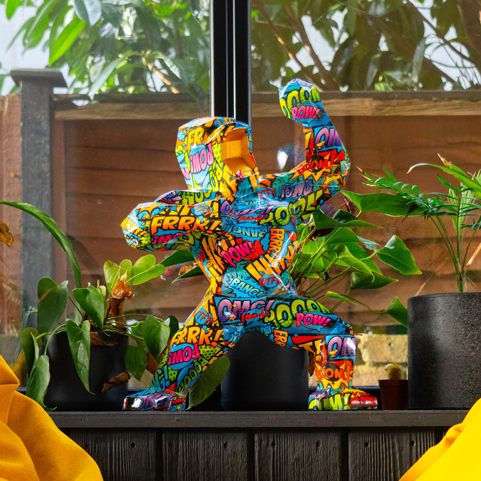 A colorful abstract sculpture of a human figure covered in comic book style text like POW! and BAM! displayed on a windowsill. Surrounding it are various green potted plants and yellow cushions, with a wooden fence visible outside.