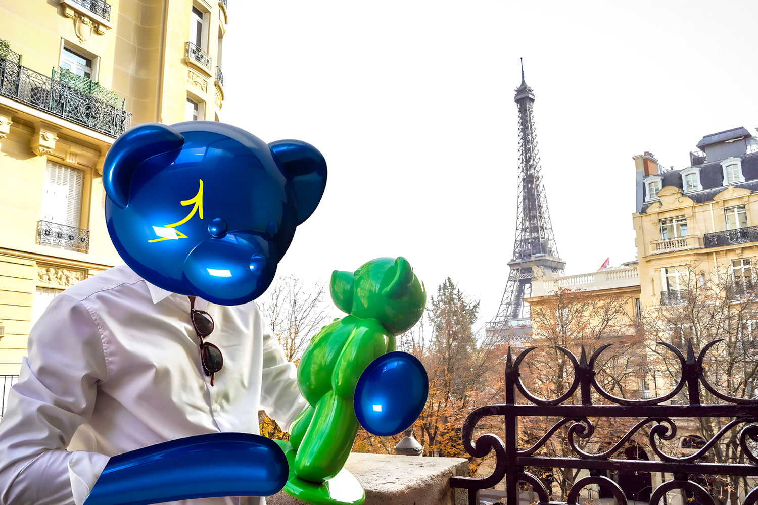 A person wearing a blue balloon bear mask holds a green balloon bear figure on a balcony. The Eiffel Tower is visible in the background, framed by surrounding Parisian buildings. The person is wearing a white shirt.