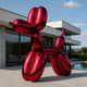 A 240cm Fire Red Balloon Dog Sculpture by Giant Sculptures stands outside a modern glass-walled house. The clear sky frames this striking art piece amidst a patio and lush green bushes.