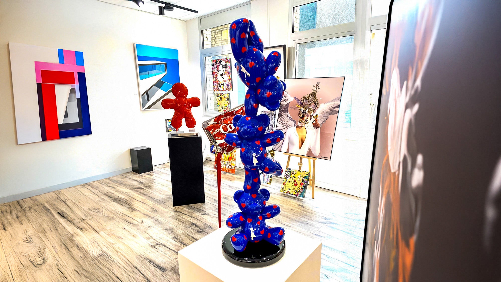 A modern art gallery with abstract sculptures and paintings. In the foreground, a blue sculpture with red patterns stands on a black pedestal. Colorful paintings and a smaller red sculpture are displayed in the background. Bright lighting fills the room.