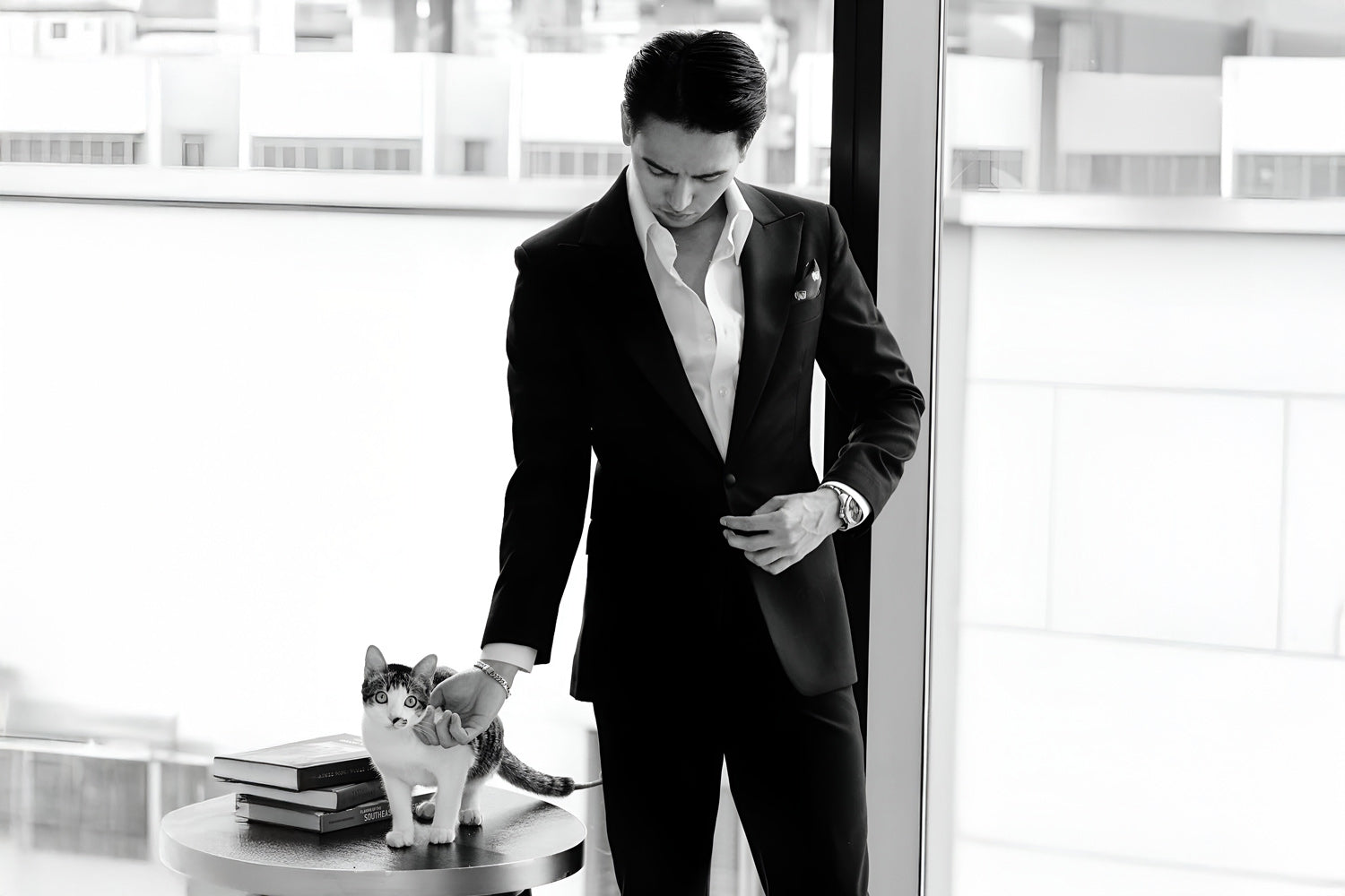 A man in a suit stands by a large window, buttoning his jacket. A cat is on a small table beside him, with a couple of books. The room has a bright, modern interior.