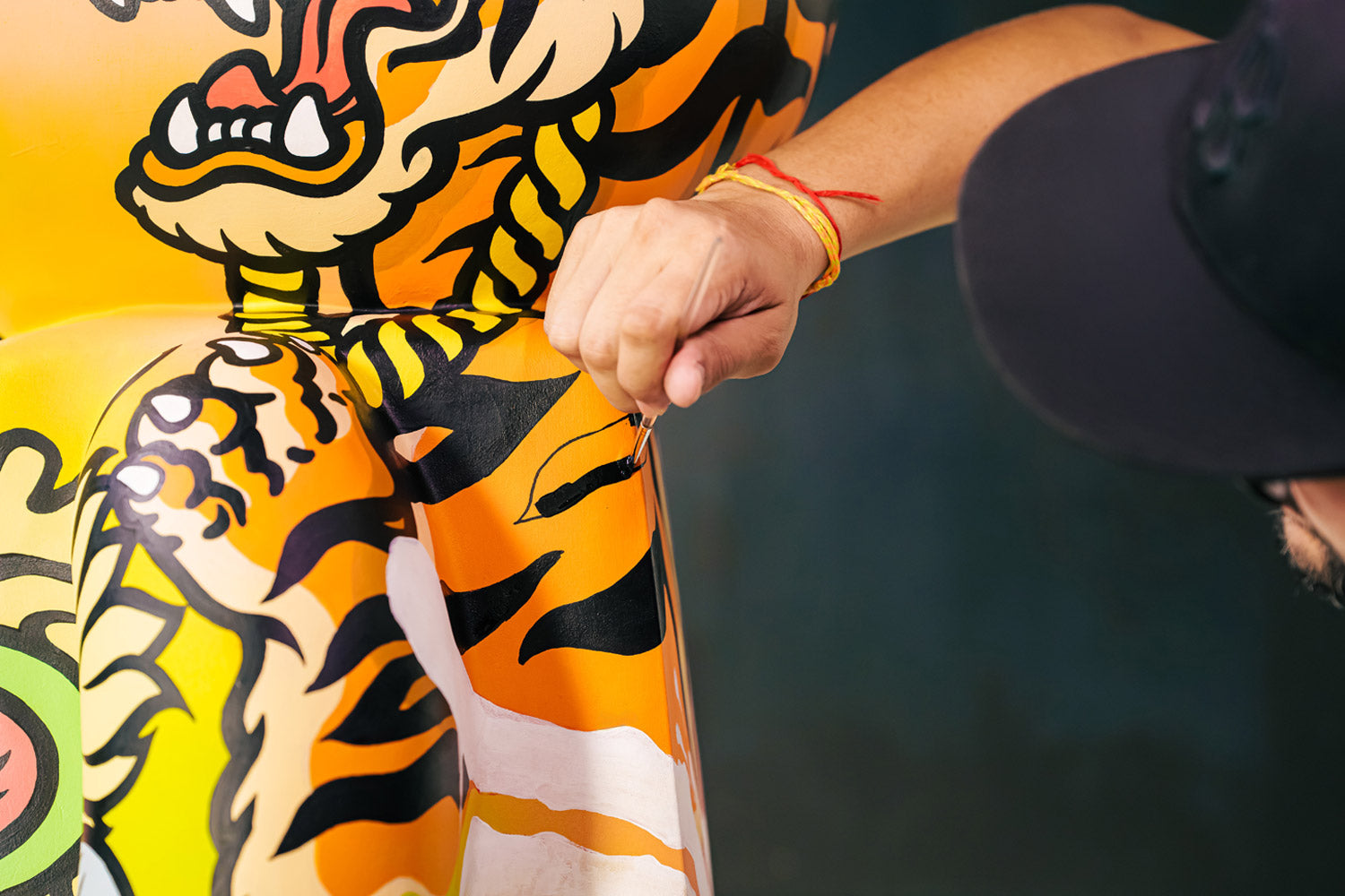 A person wearing a black cap and yellow wristbands uses a pin to puncture an inflatable object decorated with a colorful tiger design.
