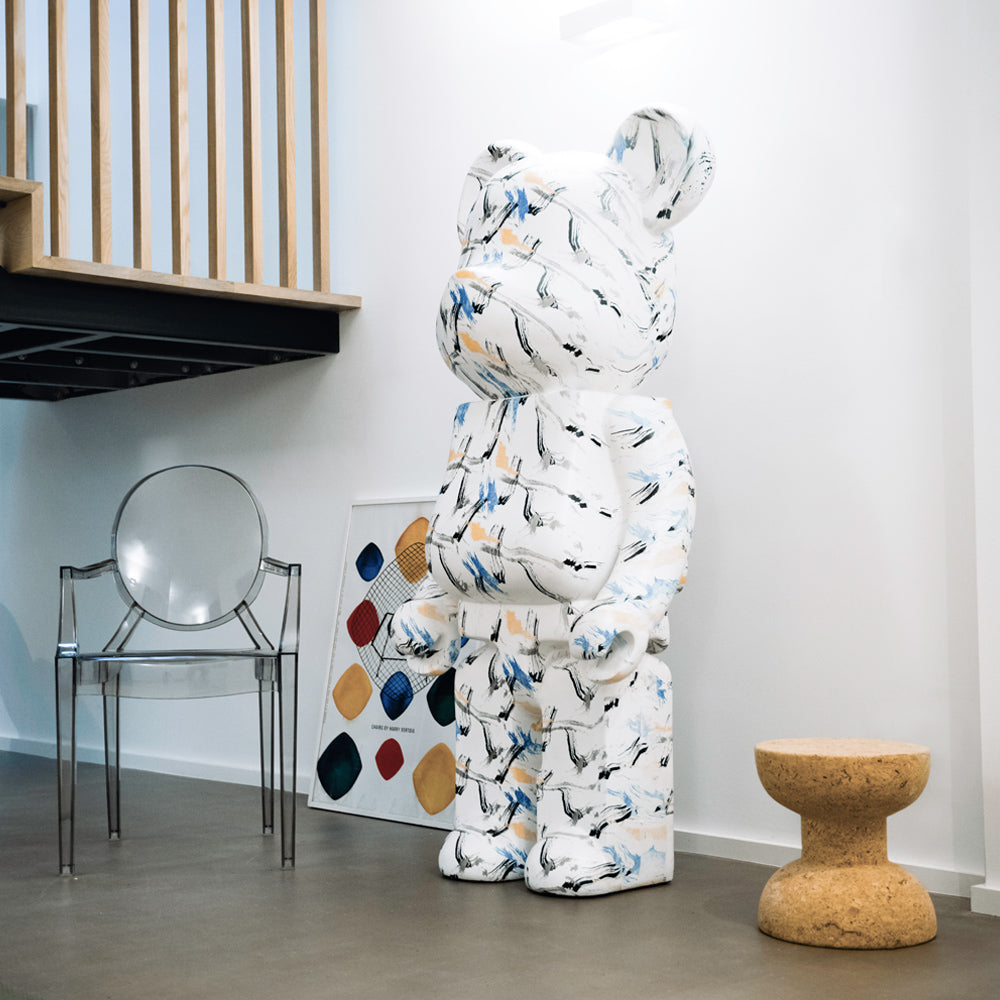 A large, abstract bear sculpture with a white background and colorful brush strokes stands in a modern room. Nearby are a transparent chair, a small cork stool, and a painting with various colored shapes leaning against a white wall.