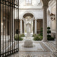 The Prayerful Angel Marble Outdoor Sculpture (220cm) by Giant Sculptures stands in an ornate courtyard with classical columns, manicured plants, mosaic tile flooring, and an open wrought-iron gate in the foreground.