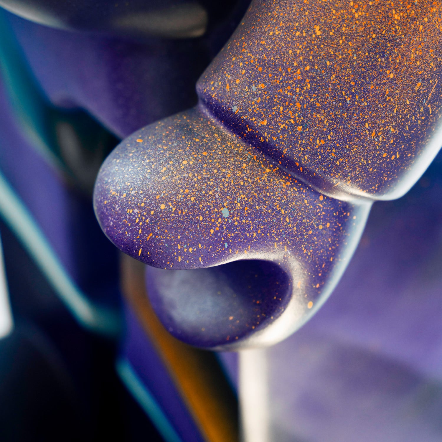 Close-up of a sculpture with a smooth, purple surface speckled with orange dots. The abstract form features a curved, loop-like shape in focus, while the background is blurred, displaying hints of blue and black.