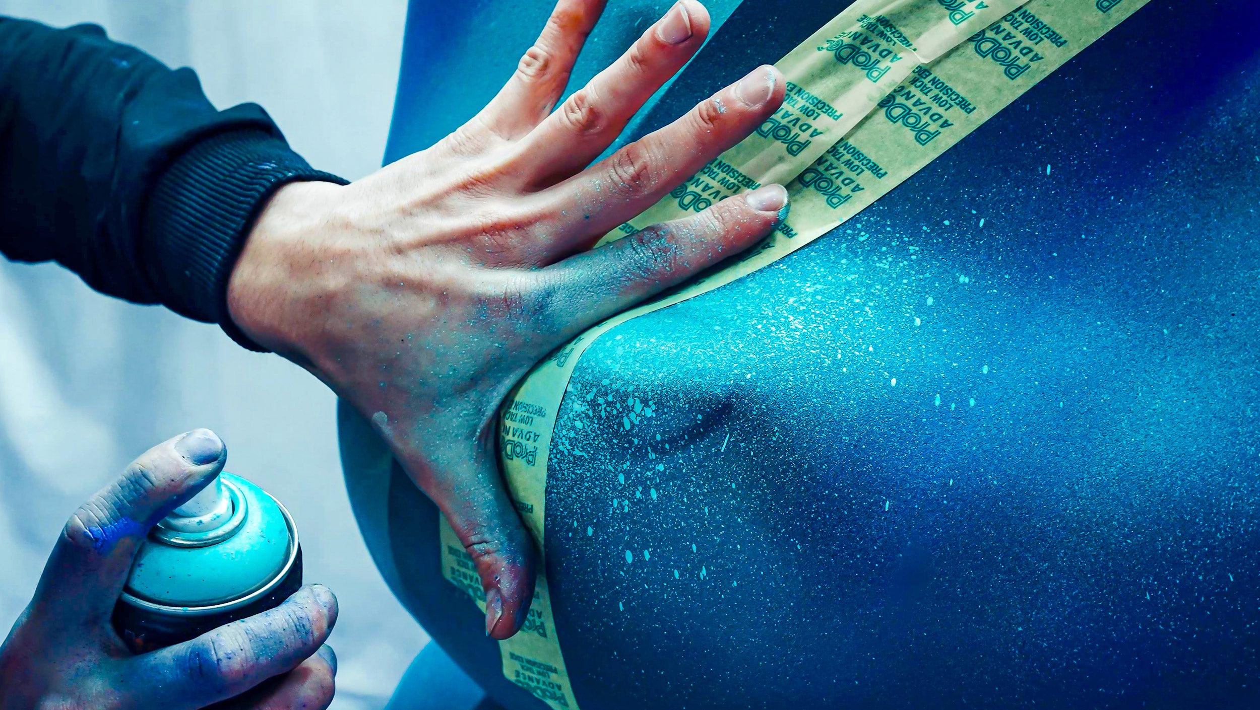 A person sprays turquoise paint onto a surface while their hand holds down a strip of tape. The hand is slightly dusted with paint, and a can of spray paint is visible in the other hand.