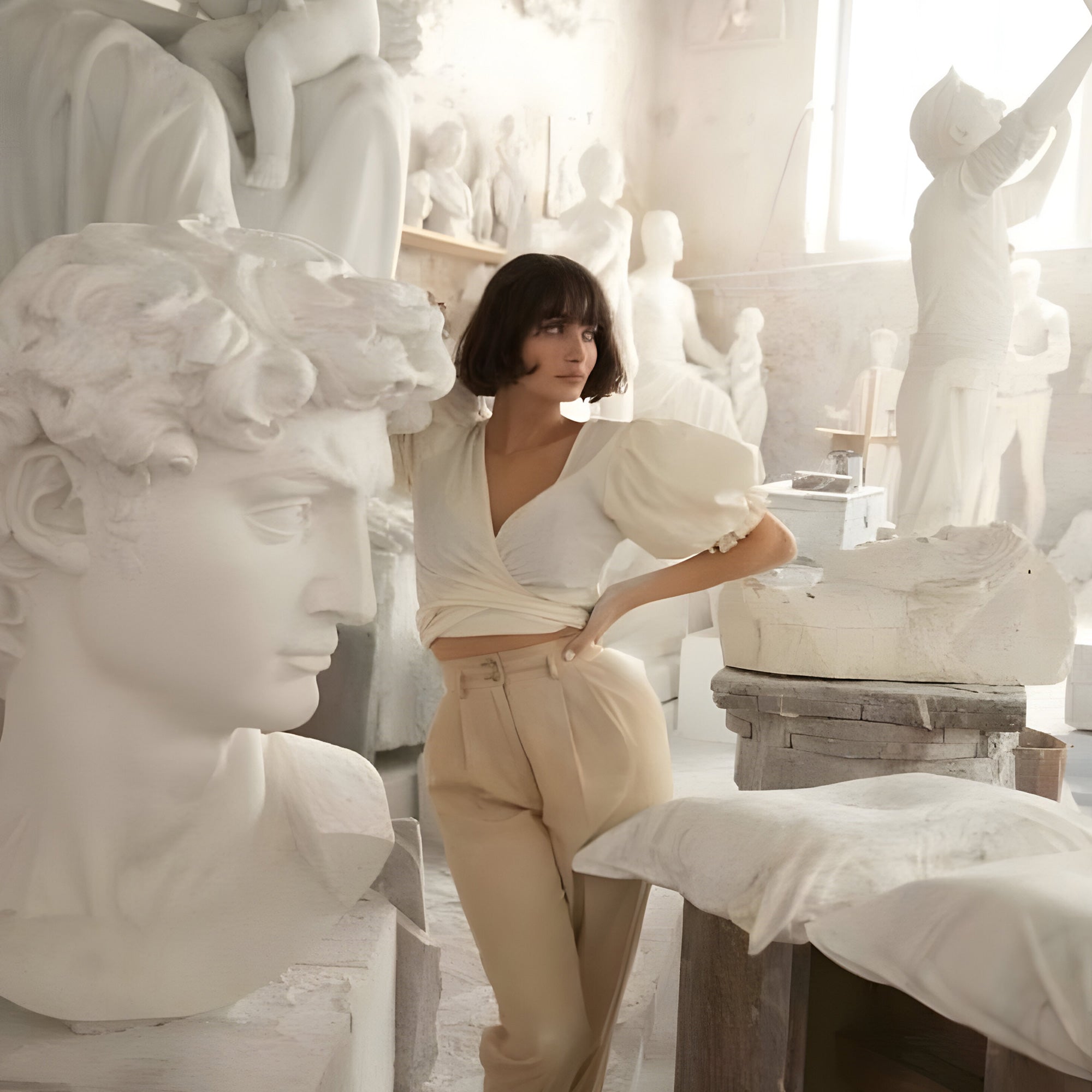 A person with a bob haircut and wearing a white blouse leans against a large classical sculpture of a head in an art studio filled with various white sculptures. Soft light streams in from a window, casting a serene atmosphere.