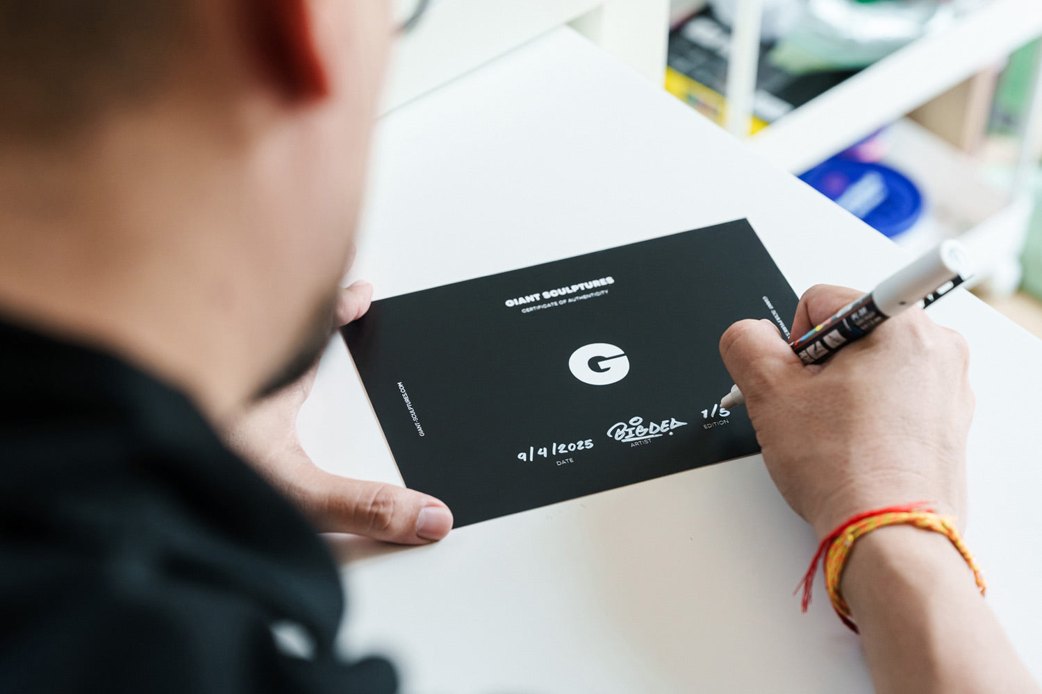 A person writes on a black card with a marker at a white table. The card displays a large G, the text GIANT GODFATHER, small white text, and decorative drawings. The persons face is not visible.