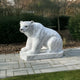 The Seated Polar Bear White Marble Outdoor Sculpture by Giant Sculptures sits on a stone pathway amid grass and bushes, showcasing a realistic smooth texture with subtle gray veining. Lush greenery and trees offer a tranquil backdrop for this stunning 50cm piece.