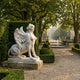 Giant Sculptures' 120cm Winged Sphinx Marble Sculpture on a weathered stone base in a formal garden, with trimmed hedges, tree-lined path, and fountain beyond.