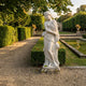 Goddess of the Harvest 180cm white marble sculpture by Giant Sculptures, holding wheat with lion head detail, on a plinth beside a formal garden pond.