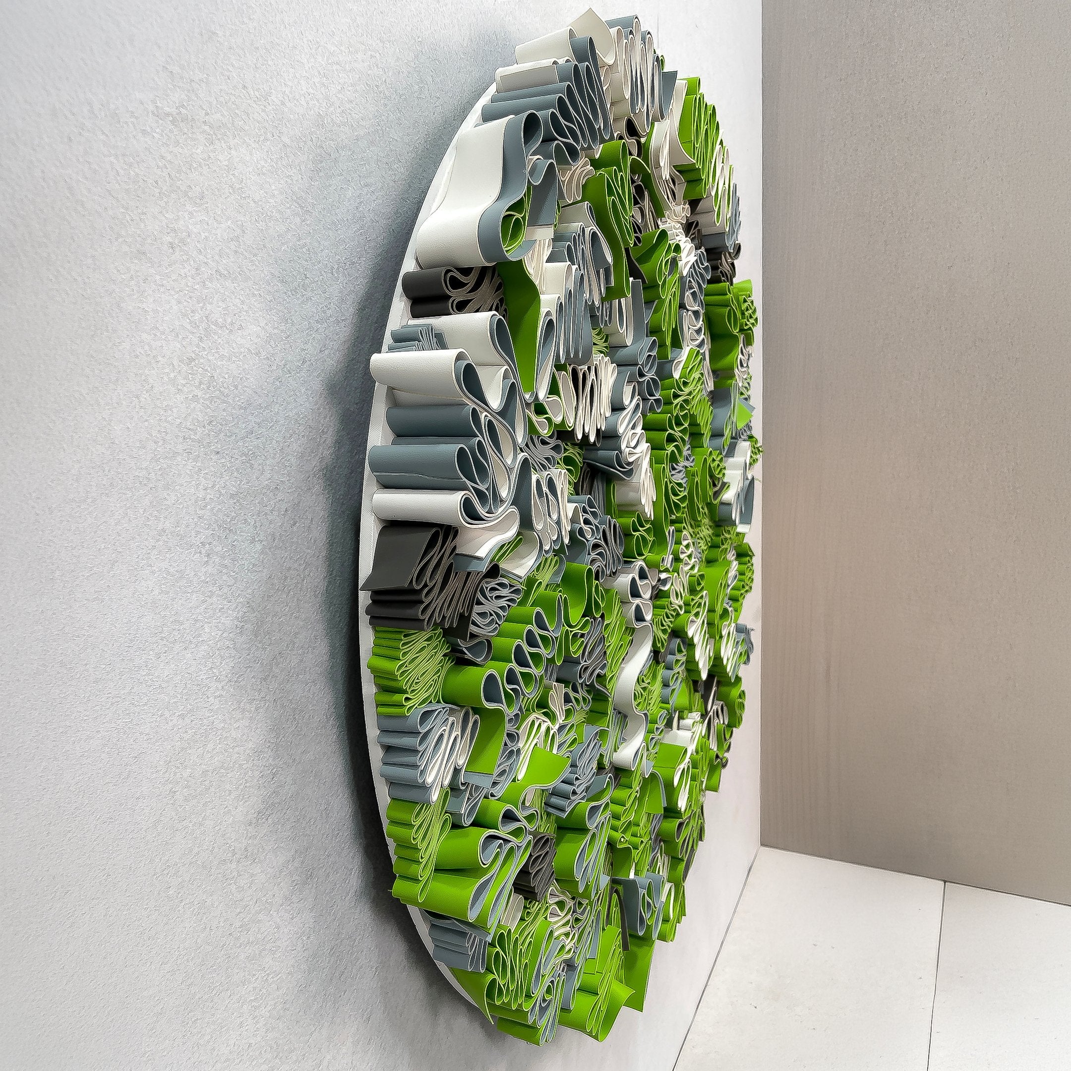 Side-angle view of the Rivora Green Ribbon Leather Circular 3D Wall Art by Giant Sculptures, showing dense green, grey, and white ribbon layers on a pale wall.