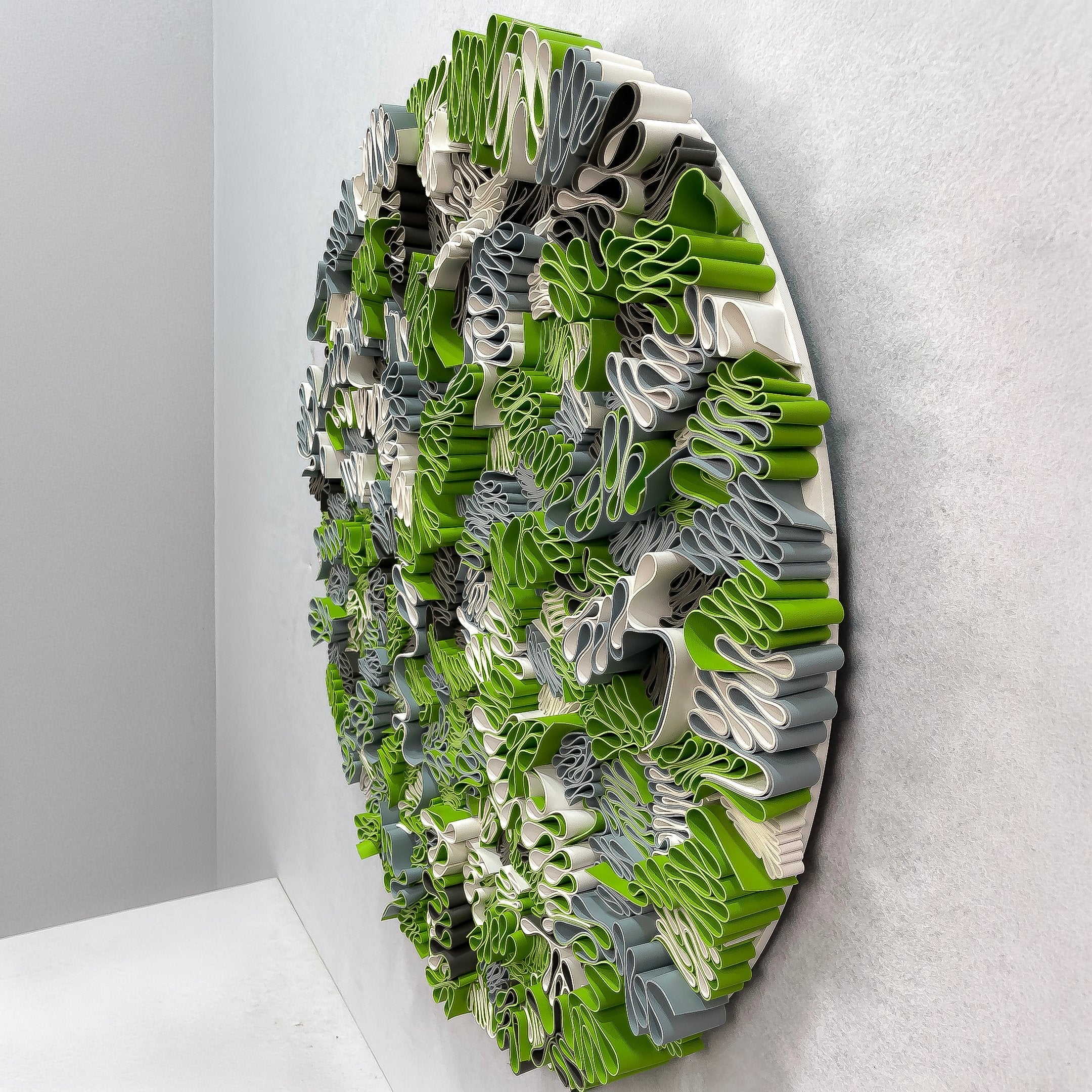 Side-angle view of the Rivora Green Ribbon Leather Circular 3D Wall Art by Giant Sculptures, showing dense green, grey, and cream ribbons mounted on a light wall.