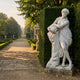 White marble sculpture of a draped female figure holding a vessel, displayed beside a tall hedge along a gravel path in a formal garden with topiary and urns.
