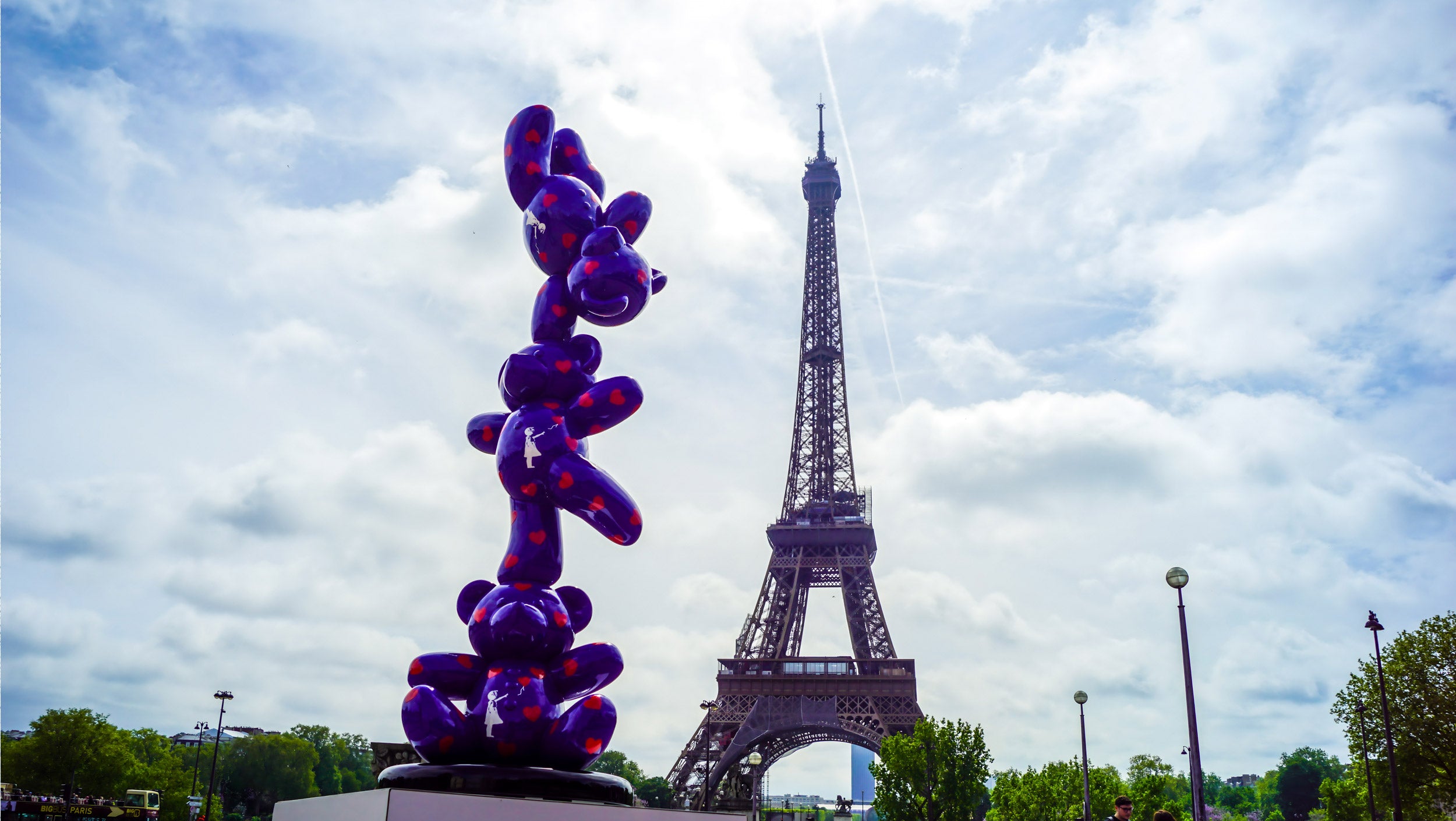 A whimsical purple sculpture resembling stacked balloons stands prominently with the Eiffel Tower in the background. The sky is partly cloudy, adding a vibrant contrast to the scene.