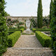 At the center of a formal garden stands the Graceful Crossed-Arms Angel Marble Outdoor Sculpture (200cm) by Giant Sculptures, surrounded by hedges, gravel paths, cypress trees, and rose-covered walls.