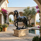 The Graceful Horse Black Marble Outdoor Sculpture by Giant Sculptures (220cm) stands on a stone pedestal in a Mediterranean courtyard with potted plants, arched walkways, and a small pool, overlooking the distant sea.