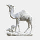 The Classic Caravan Camel Marble Outdoor Sculpture by Giant Sculptures is a 160cm white marble piece featuring an intricately detailed standing camel with a decorated saddle rug on a rocky base, its head slightly raised, and a small bush at its feet, showcasing traditional craftsmanship.