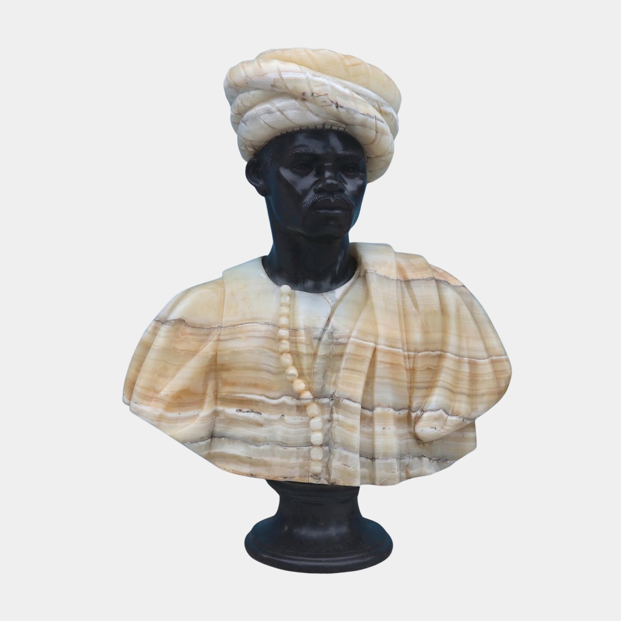The Turban Marble Bust Sculpture - 75cm by Giant Sculptures features a two-tone marble portrait of a man in a turban and robe, accented with a beaded necklace and mounted on a pedestal, crafted in contrasting light and dark stone.