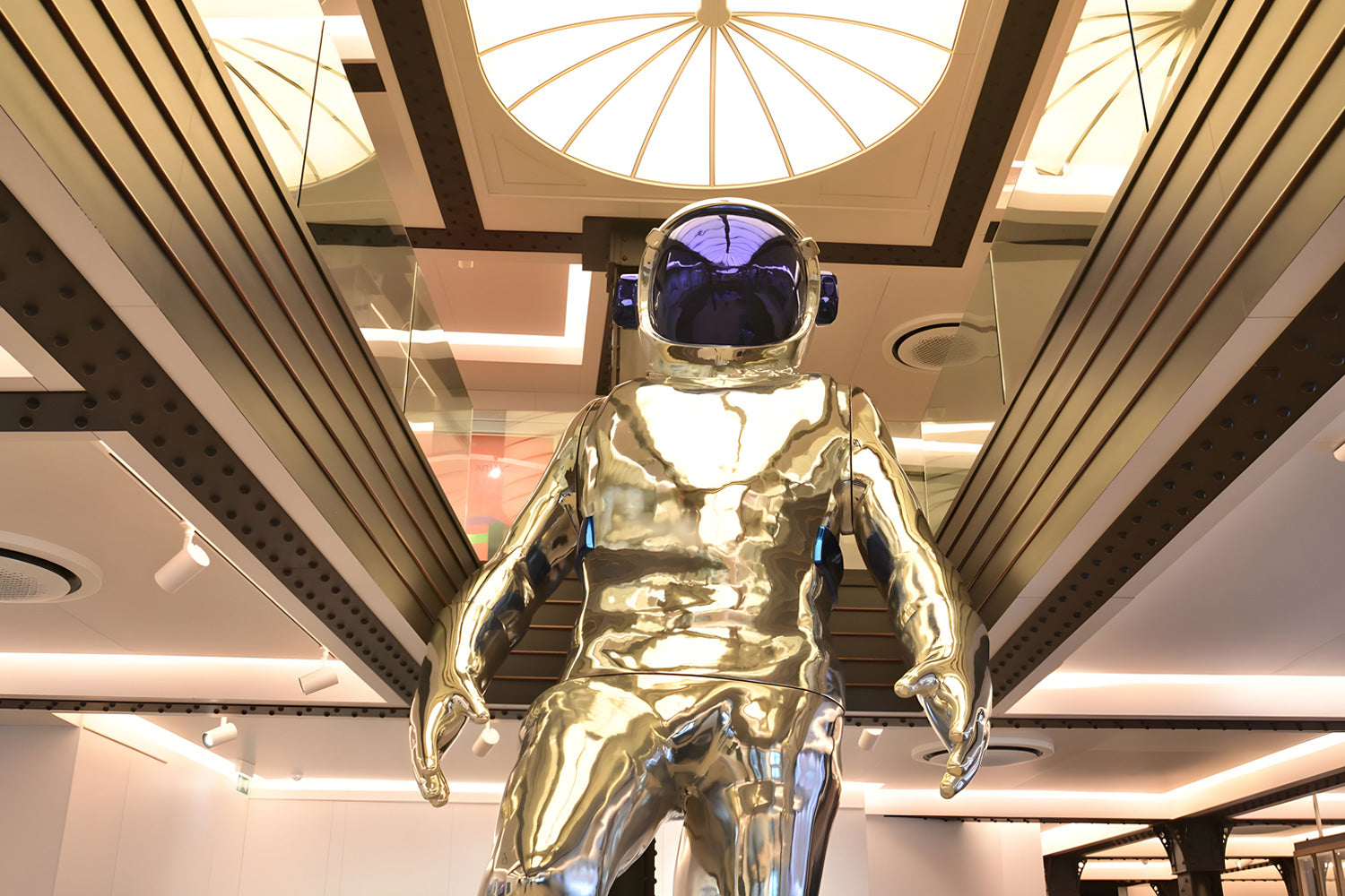 A reflective, silver astronaut statue stands prominently indoors, viewed from a low angle. The figure wears a helmet with a blue visor. The ceiling features a bright, round skylight and modern architectural details.