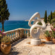 Giant Sculptures' 100cm Abstract Knot Marble Sculpture in white marble on a coastal stone terrace with balustrade, potted plants, cypress trees, and sea view.