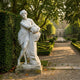 White marble sculpture of a draped female figure holding an urn, displayed on a stone plinth beside a hedge in a formal garden with topiary and fountain.