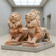 The Giant Sculptures Giant Lion II Yellow Marble Outdoor Sculptures (180cm) stand on rectangular bases in a minimalist modern gallery with neutral walls and a high skylit ceiling.