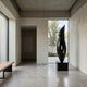 Abstract Flame Marble Sculpture in black marble on a black plinth, displayed in a minimalist concrete hallway beside a window framing a birch tree.