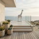 A modern terrace with potted plants overlooks the ocean. The Regal Giraffe White Marble Outdoor Sculpture (350cm) by Giant Sculptures stands on the tiled platform near glass railings, complementing the serene coastal landscape.