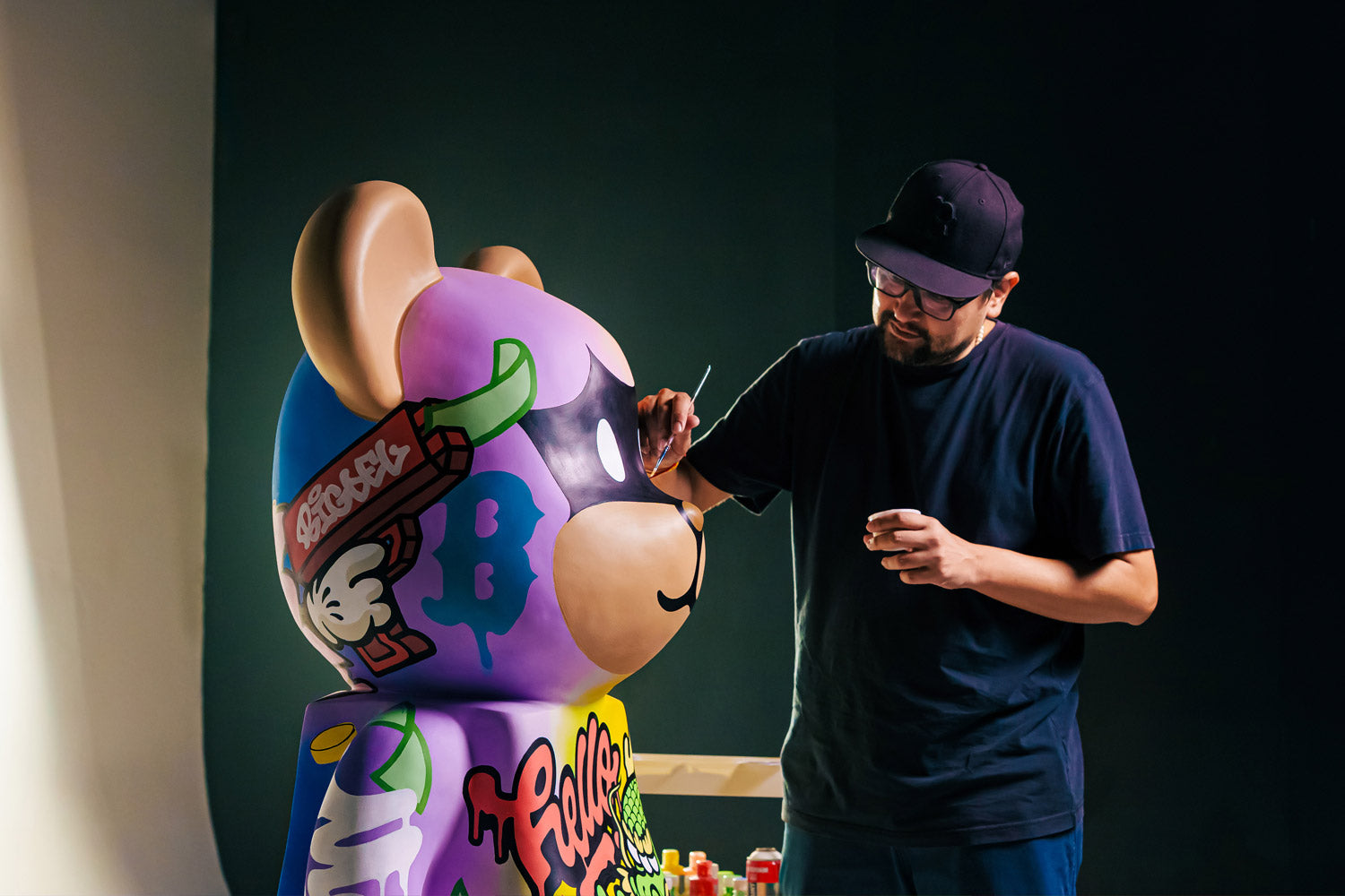 A person wearing glasses and a cap paints colorful graffiti-style designs on a large bear-shaped sculpture in a studio setting.