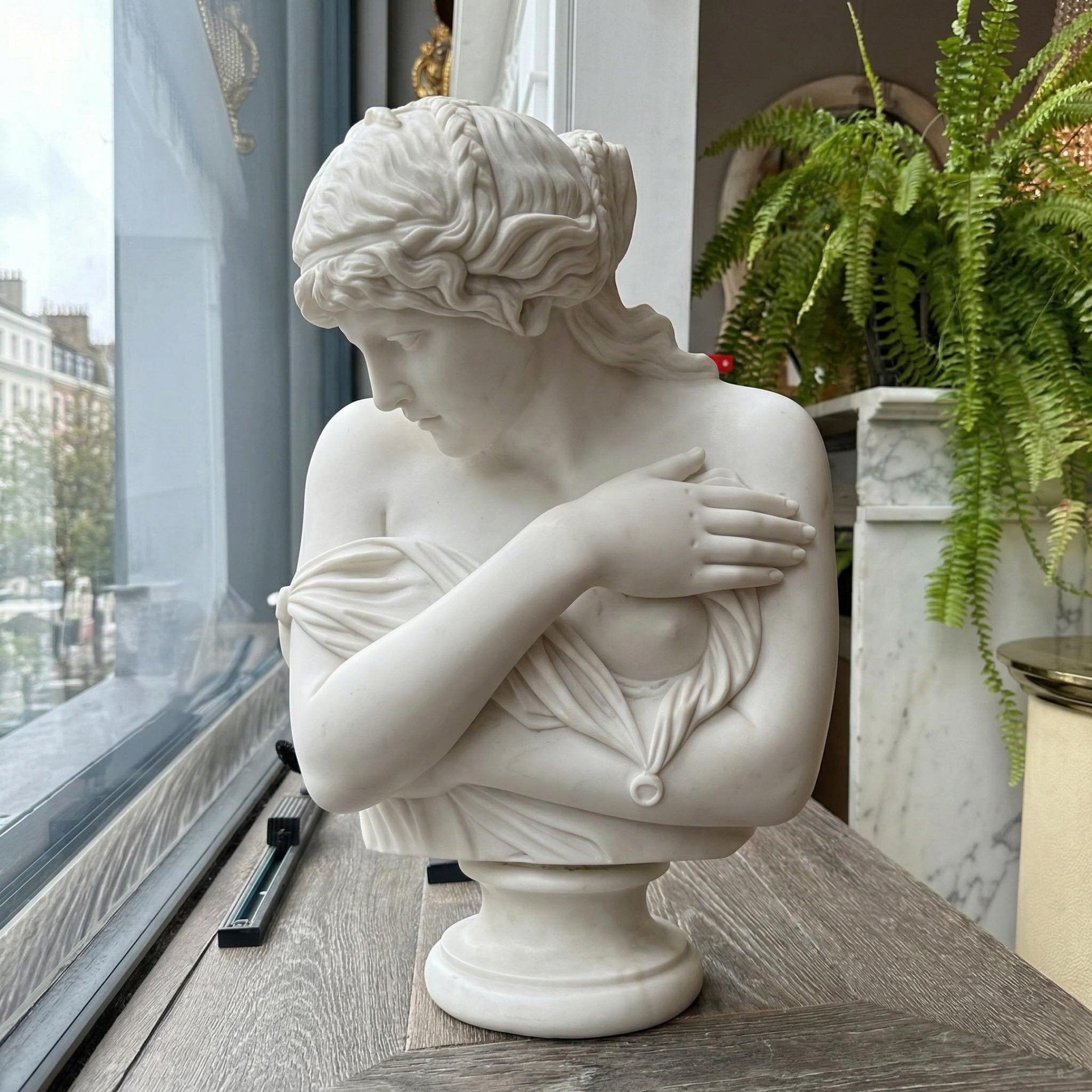 The Psyche Marble Bust Sculpture - 65cm by Giant Sculptures features a woman with wavy hair, gazing downward with her right arm across her chest. This classical piece sits on a wooden surface near a window, surrounded by plants and city views.