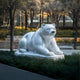 The Resting Polar Bear White Marble Outdoor Sculpture by Giant Sculptures, at 65cm, sits serenely in a garden amid trees, atop a low pedestal. Surrounded by modern buildings and soft lighting, it creates an enchanting outdoor display.