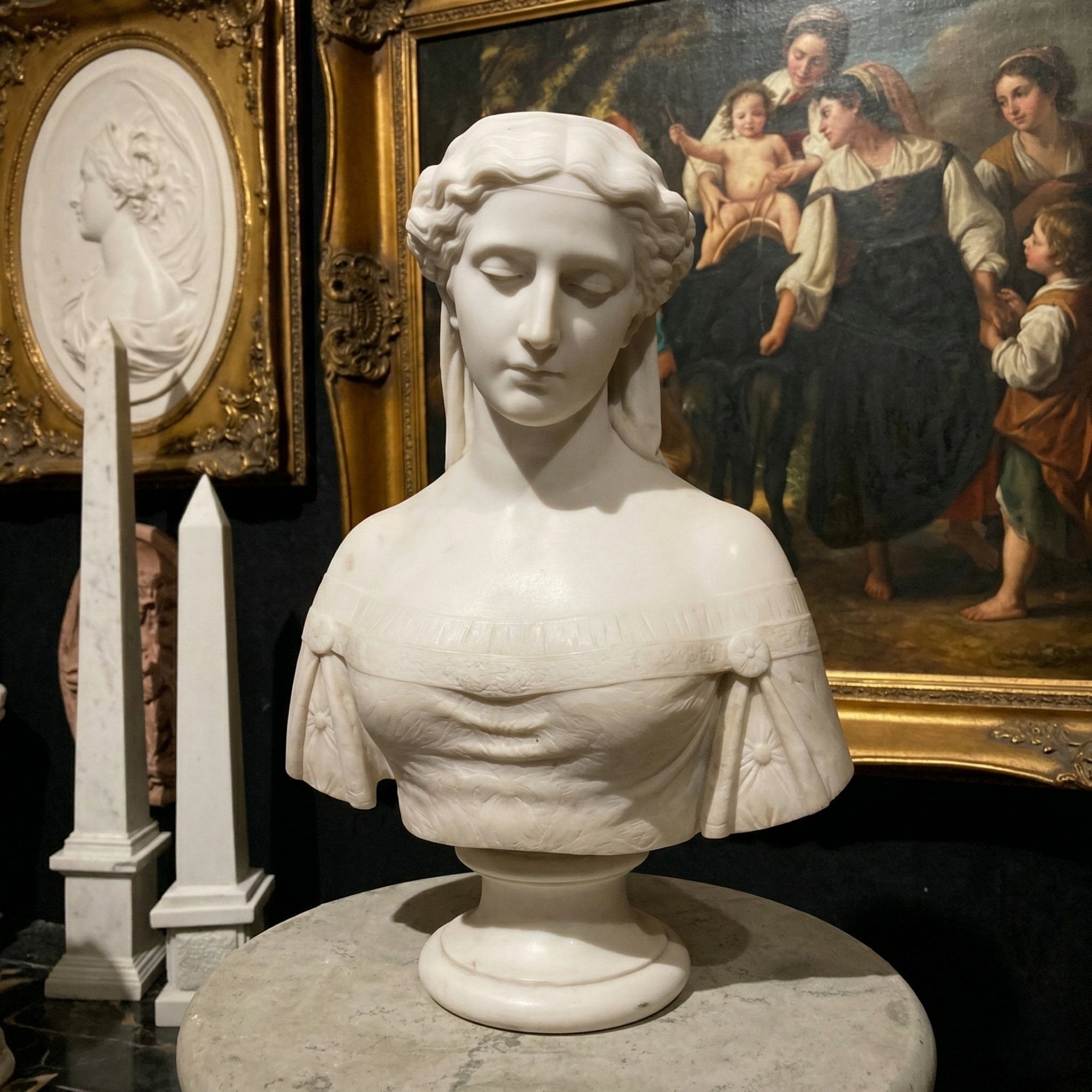 The Flora Marble Bust Sculpture - 65cm by Giant Sculptures, a serene female figure in natural white marble with wavy hair, is showcased on a pedestal among framed artworks and sculptures in an art gallery.