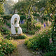Giant Sculptures' 80cm Abstract Intertwined Forms Marble Sculpture in white, displayed on a stone base in a cottage garden with foxgloves, delphiniums, and roses.
