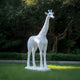 The Regal Giraffe White Marble Outdoor Sculpture by Giant Sculptures stands 350cm tall on a grass lawn amidst trees and bushes. This hand-carved piece features a textured surface, offering a timeless wildlife presence in the serene garden setting.