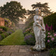 Venus Bathing Marble Sculpture 160cm by Giant Sculptures, standing on a stone path in a walled garden with roses, lavender, and foxgloves at dusk.