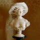 The Lady With Veiled Hat Marble Bust Sculpture - 65cm by Giant Sculptures is intricately crafted to showcase the delicate veil folds, set against a backdrop of patterned golden wallpaper.
