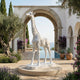 The Graceful Giraffe White Marble Outdoor Sculpture - 350cm by Giant Sculptures stands on a circular platform in a Mediterranean courtyard with arched architecture, potted plants, lush greenery, and a sea view—a stunning garden feature.
