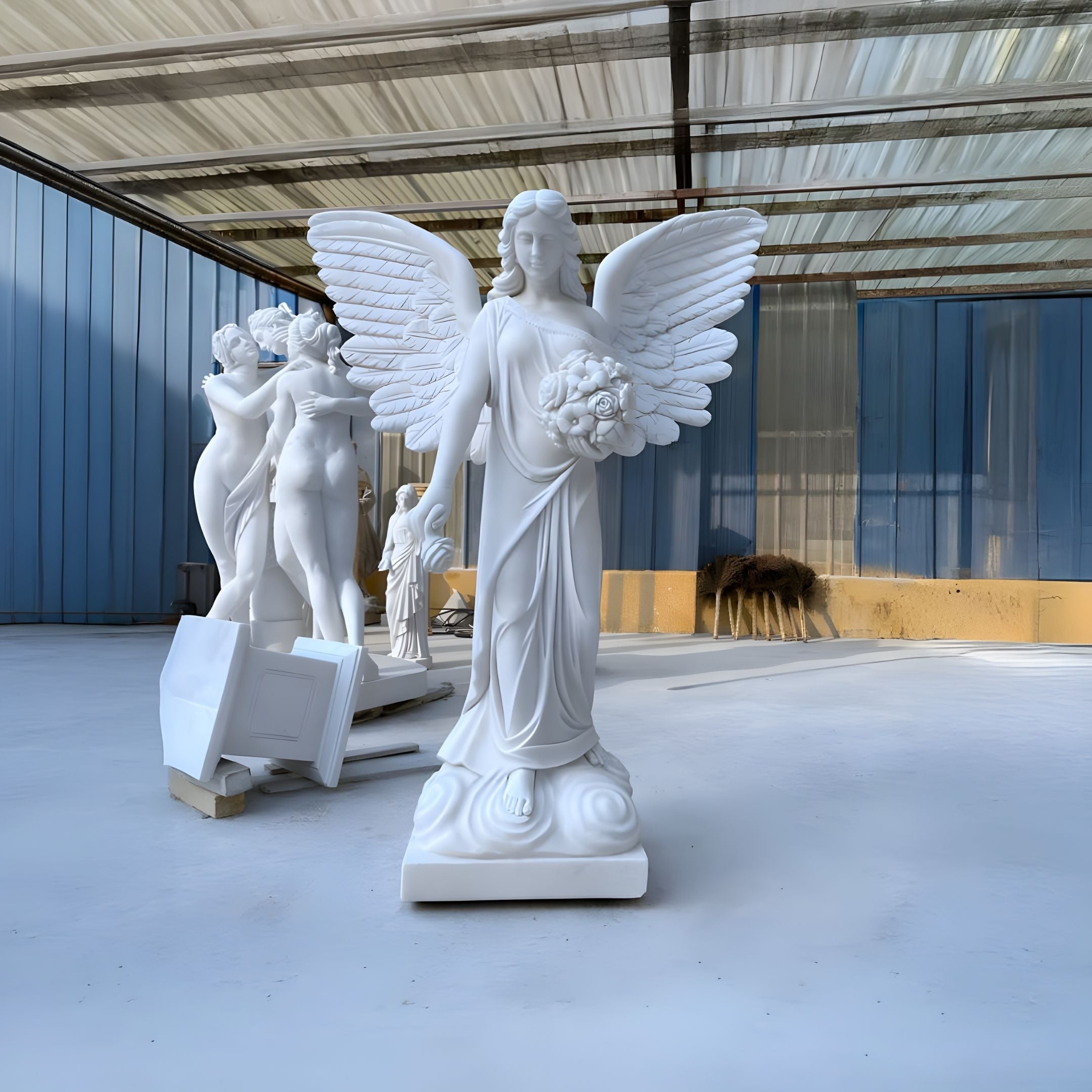 A spacious area features Giant Sculptures Winged Flower Angel Marble Outdoor Sculpture (200cm) in white, holding a bouquet. Nearby, two marble sculptures of an embracing couple are partially shadowed against blue and beige walls.