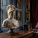 The Athena Marble Bust Sculpture (90cm) by Giant Sculptures stands on a wooden desk amid dark blue walls, framed portraits, red curtains, and antique books, evoking the ambiance of a classic marble collection.