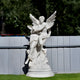 The Angelic Embrace Marble Outdoor Sculpture by Giant Sculptures, standing at 260cm, features a white marble angel embracing a person on a round base. Set against a gray fence, it beautifully contrasts the lush greenery in the background.