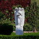 The Blooming Guardian Angel Marble Outdoor Sculpture by Giant Sculptures stands gracefully amid lush greenery. This 220cm white sculpture holds flowers surrounded by vibrant red and green foliage, creating a serene, tranquil garden decor scene.