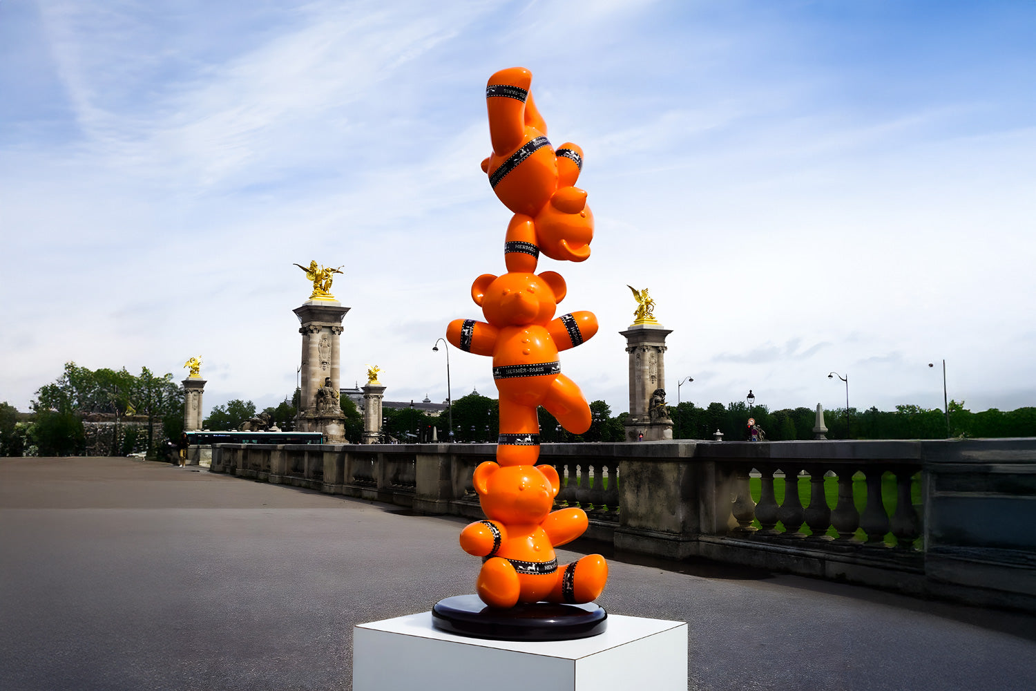 A sculpture of three stacked orange bears with black bands stands on a pedestal outdoors. The background features an ornate bridge with golden statues and a cloudy sky.