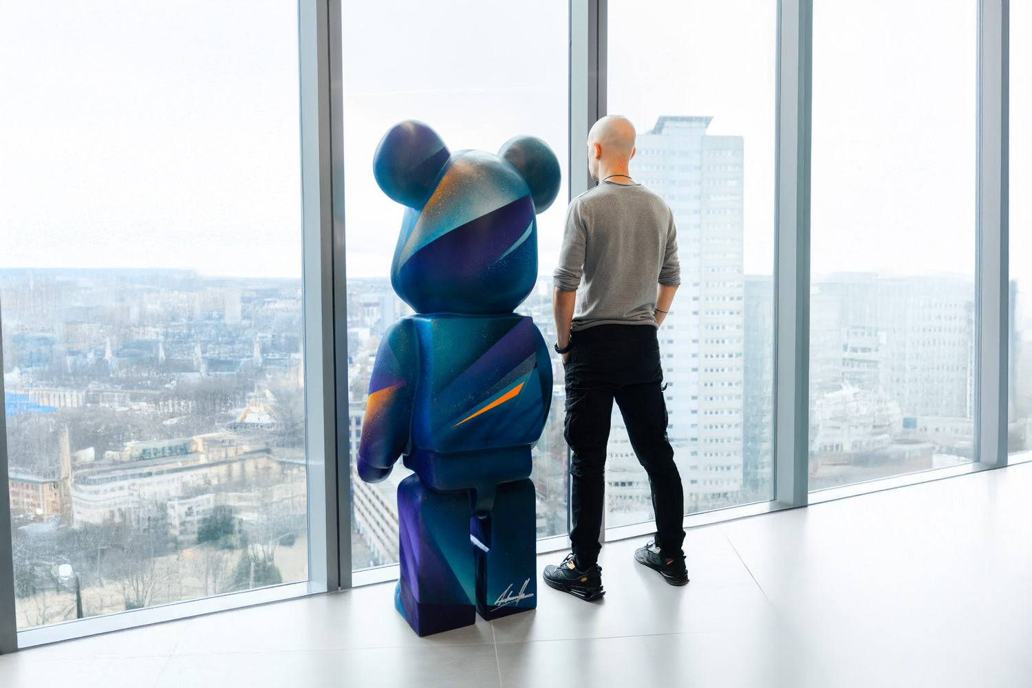 A person stands next to a large, colorful bear-shaped sculpture, both overlooking a cityscape through floor-to-ceiling windows in a modern high-rise building. The sky is overcast, casting a muted light over the scene.