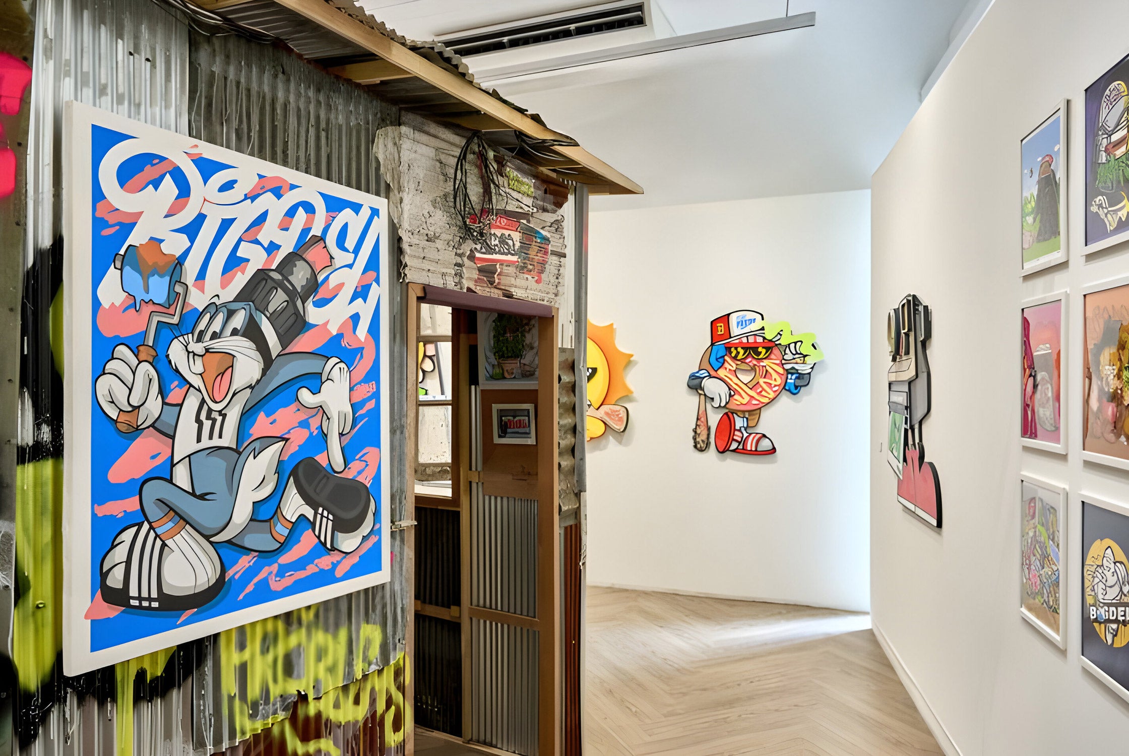 A gallery interior featuring vibrant graffiti art on white and corrugated metal walls. The artwork includes cartoon characters with exaggerated expressions and bold colors. The floor is wooden, and the space has modern lighting.